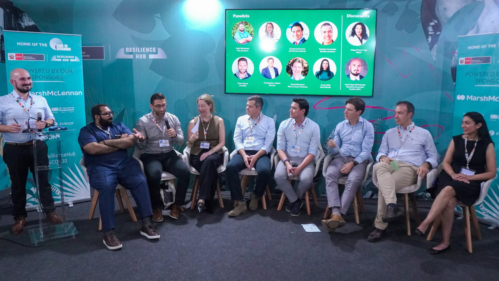 Nine people sit on chairs in a panel discussion, with microphones and badges, facing an audience. A screen behind them displays panelist photos and discussion topics.