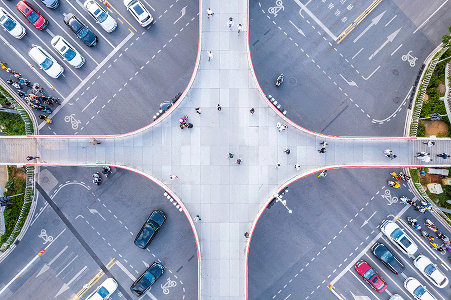 Aerial view of a cross-shaped pedestrian overpass above a busy intersection with cars and people visible on the roads and sidewalks below.